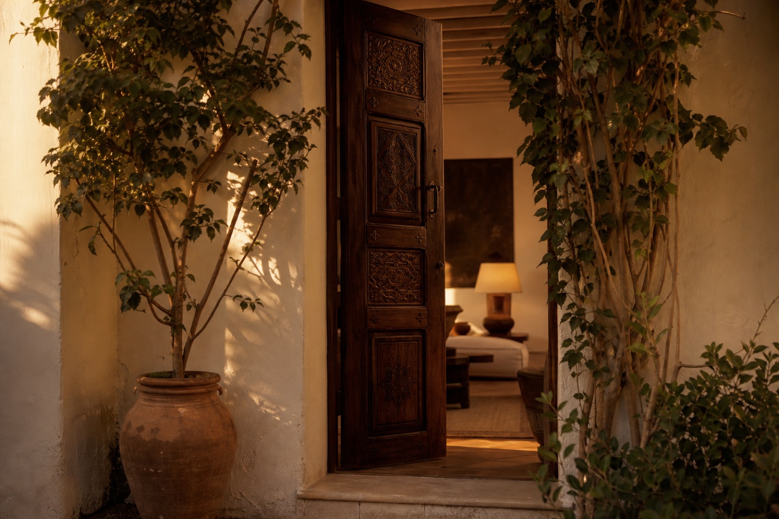An open carved wooden door between climbing plants, revealing a softly lit room.